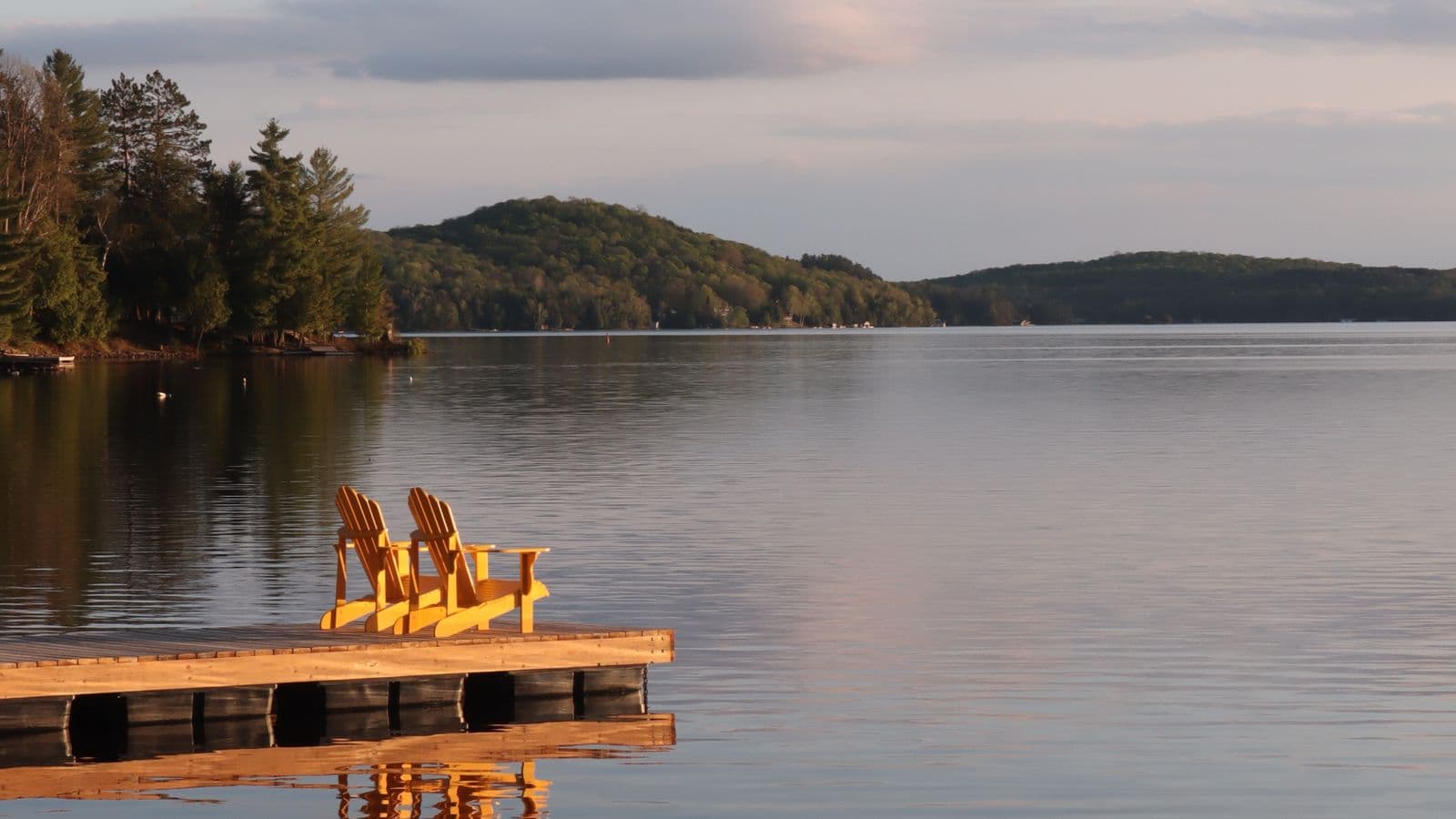 A dock with two Adirondack chairs facing a calm lake at sunset